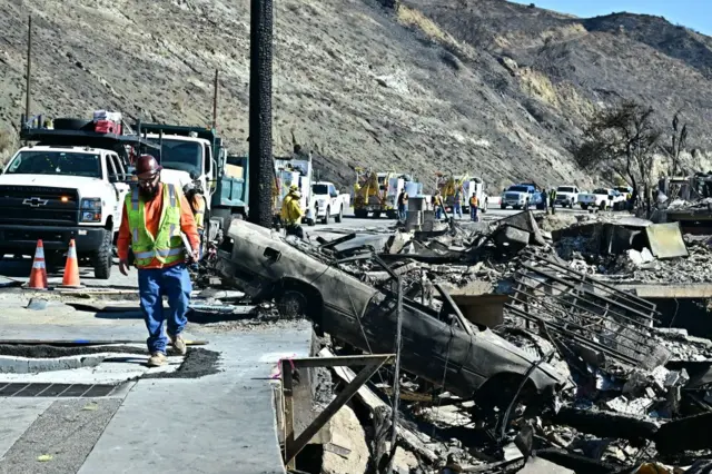 A man in a hard hat and high vis walks along a road, which has several trucks parked on it. To one side, the street is lined with burned down structures and a ruined car