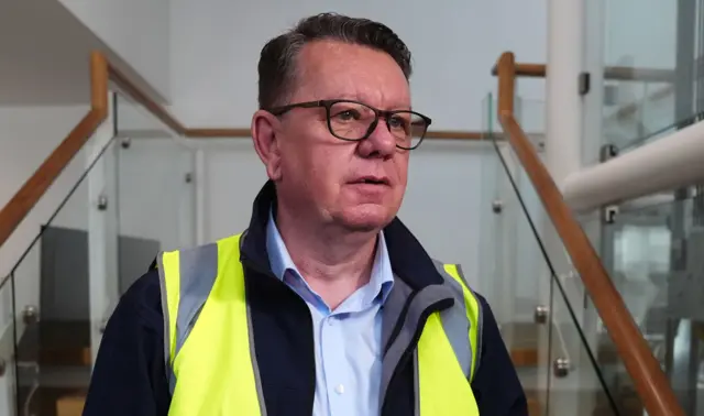 A man with dark hair and glasses wearing a hi vis jacket in front of a stairway