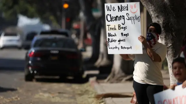 A person standing on side of the road holds a sign "We need cleaning supplies - trash bags, bleach, rags, rubber gloves, laundry detergent"