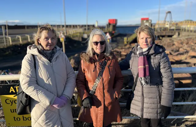 Three women standing in front of railings in front of a harbour.