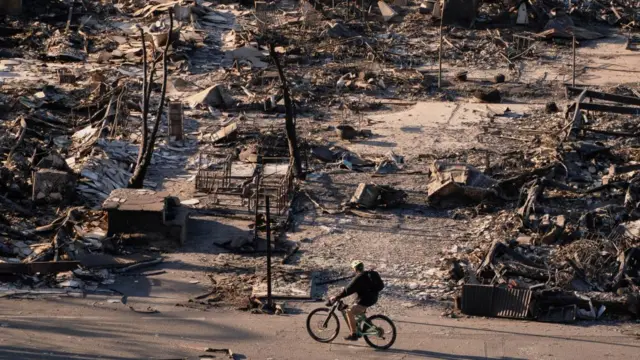 Man on a bicycle rides pas a large open space full of burnt debris from properties