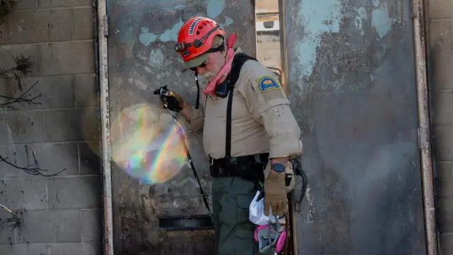 A Los Angeles County Sheriff's Department volunteer leaves a burned property searched for hazards and remains as the Eaton Fire continues, in Altadena, California, U.S. January 12, 2025. He is wearing a red helmet and beige uniform top with khaki cargo trousers.