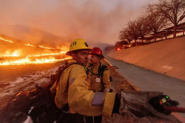 Two firefighters in yellow suits stand in front of a blazing fire