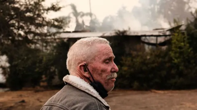 Headshot taken from side on of Rich standing in front of a smoking, burnt structure