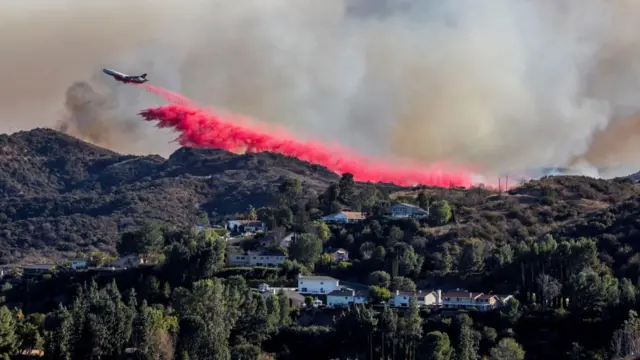 A plane flies low over hills, leaving a trail of bright pink substance behind it