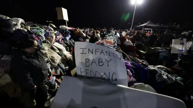 Piles of clothing and bedding with a hand-written sign saying Infants/Baby bedding at the Santa Anita Park in Arcadia