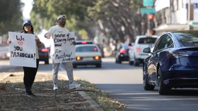 Two people stand on the roadside with signs asking for cleaning supplies and another sign saying "We love you Pasadena"