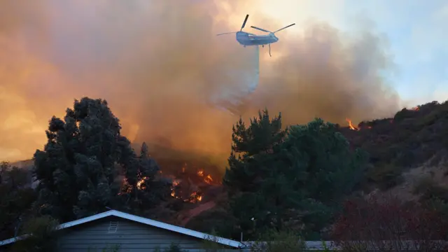 A helicopter does a water drop over a home during the Palisades wildfire in Los Angeles