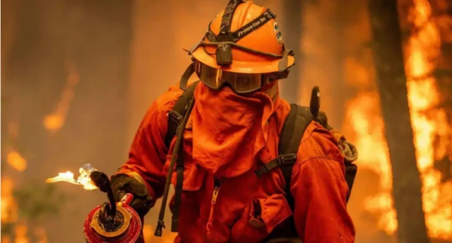 An incarcerated firefighter wearing an orange hasmat suit and goggles, in front of a blazing orange fire