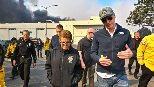 California governor Gavin Newsom (right) and LA mayor Karen Bass visit areas of the Palisades fire with authorities in background and dark smoke clouds behind a building