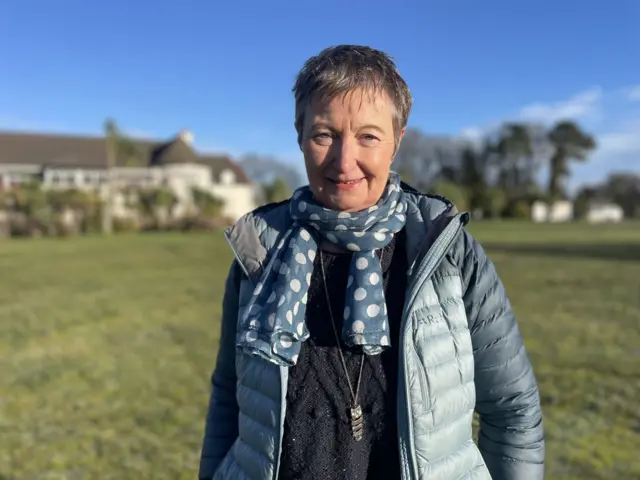 Woman with short hair wearing a blue padded jacket and blue and white polka dot scarf standing outdoors with large building slightly visible in the far distance