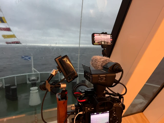 Digital camera on a tripod with microphone, lighting and iPhone attached, positioned looking out over the water with flags showing and a grey sky and sea