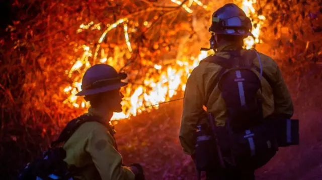 Firefighters with their backs to the camera look on at a glowing fire in woodland where the Palisades Fire is roaring