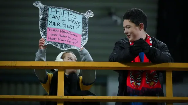 A young Dagenham & Redbridge fans holds aloft a tin foil FA Cup asking for Harry Phipps' shirt