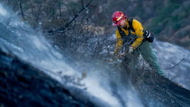 A firefighters works alongside a mountain during the daytime to extinguish smoke emanating from the ground