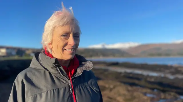 Woman smiling, wearing a grey waterproof jacket with a red jumper visible underneath, standing on a rocky beach with water in the far distance