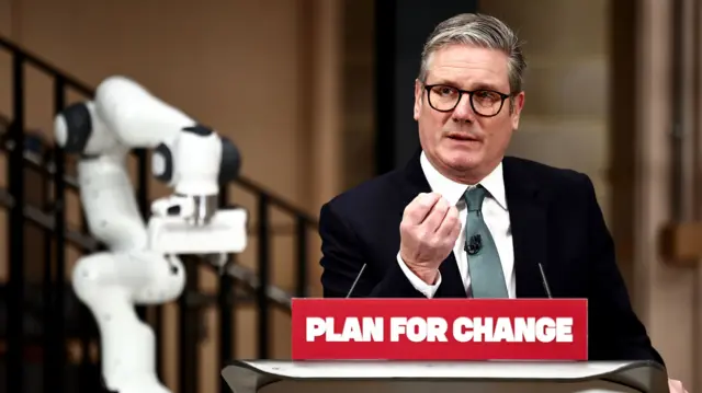 Starmer wears a suit and speaks from a podium featuring a red sign reading 'Plan for Change'. He looks to the left of the camera