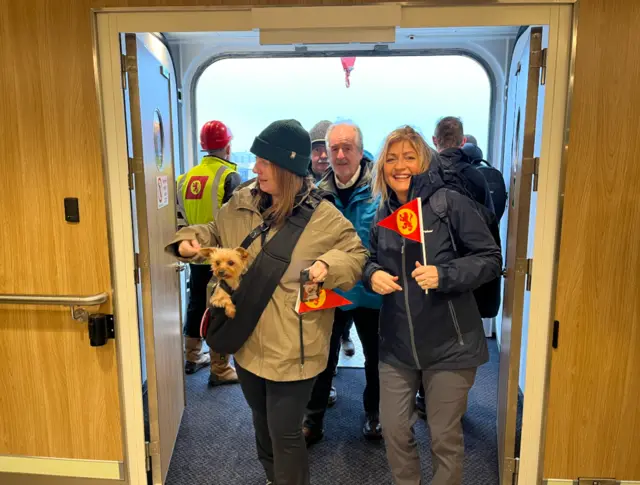 Excited passengers wave flags and carry a dog