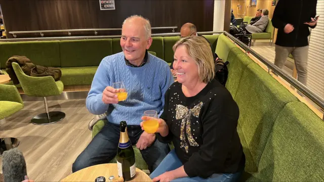 Man in a blue jumper and woman in a black jumper with a sequin bow, holding glasses with a bottle of champagne in front of them