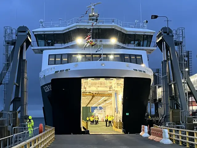 Exterior of Glen Sannox with cargo door open showing car area empty and brightly lit with crew members standing inside