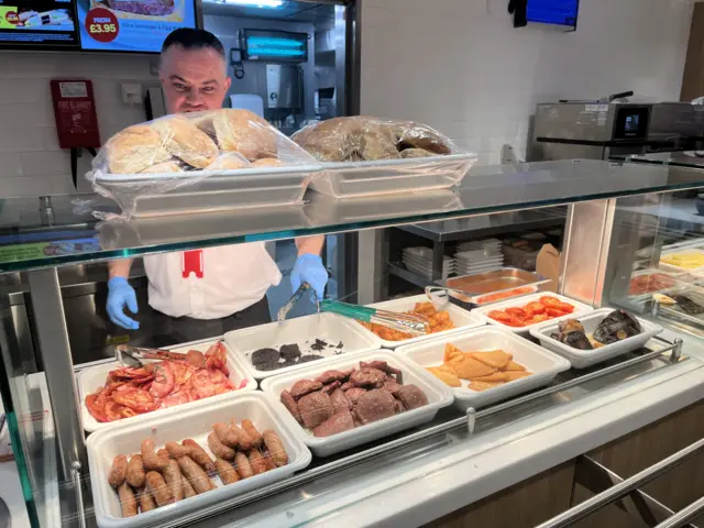 Man wearing white chef shirt and blue gloves stand behind a hot plate filled with breakfast foods below lights with bread rolls on top