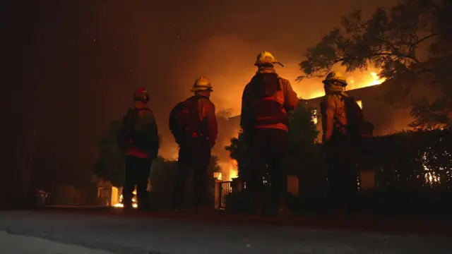 Backs to the camera, firefighters look on at a home being overcome by flames