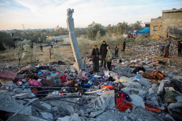 A group of people stand next to a ruined building that is covered in rubble