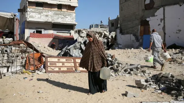 A displaced Palestinian woman, Makram Hamdouna, carries a water container outside her shelter, in Khan Younis