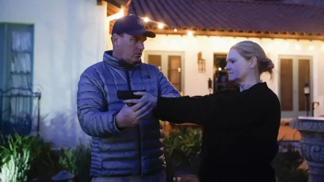 A man in a puffer jacket positions a woman's arms as she points a gun and looks ahead