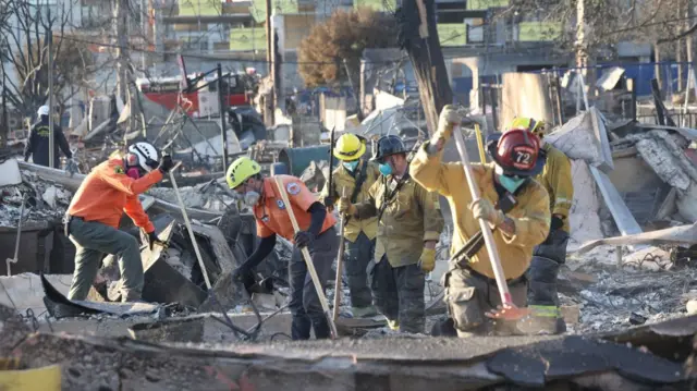 Rescue workers in orange and yellow long-sleeved polo shirts look through the debris of burned down buildings