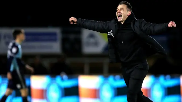 Matt Bloomfield is pictured celebrating with his arms out stretched. A Wycombe player can be seen, out of focus, in the background