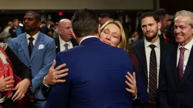 Hegseth, with his back to camera, hugs his wife, while people in the room look on