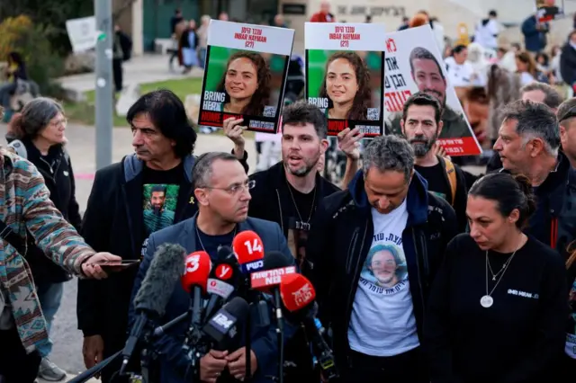 A group of people with signs asking for hostages to come home stand in front of a microphone and look amongst themselves