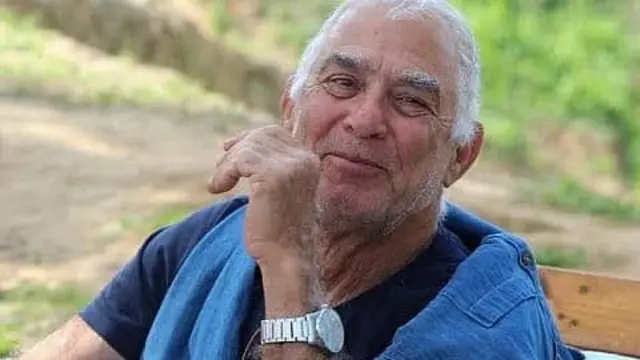 A headshot of Gadi Moses smiling to the camera, he is an older man with grey hair and stubble and wears a silver watch