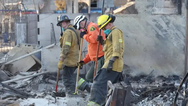 Three rescue personnel in safety gear stand with helmets at a site of a burned out building, surrounded by rubble