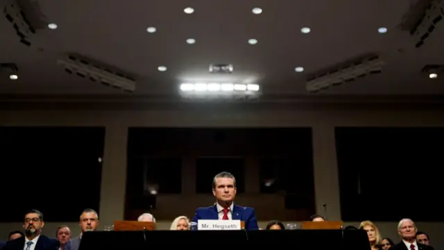 A wide shot of Hegseth sitting in the chamber, in front of his name tag