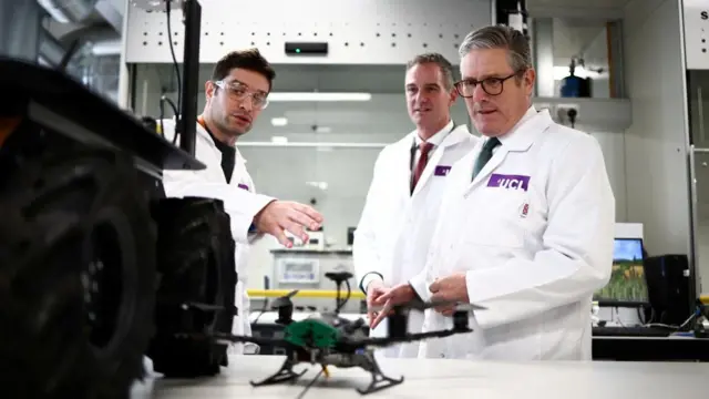 Prime Minister Keir Starmer visits the Manufacturing Futures Lab at University College London. He standing with two researchers, all wearing white coats, and looking at what appears to be a drone.