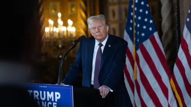 Trump standing in front of a lectern, flanked by US flags