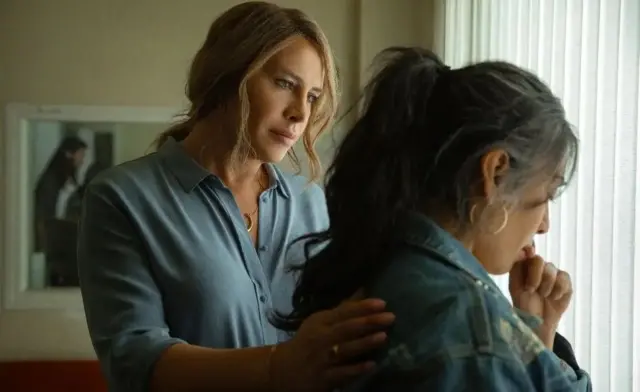 A screenshot of Emilia Pérez, showing one woman placing a hand on another's back as they stand together in a room behind a curtain