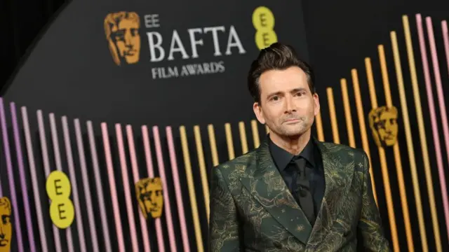 David Tennant at the Bafta Film Awards 2024. He is smartly dressed with Bafta branding behind him