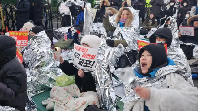 People sitting on the ground, wrapped in thermal blankets and appearing to sing. A protester is holding a phone with its flashlight on.