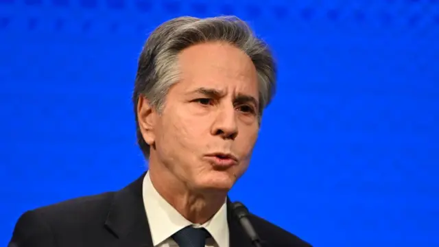 Antony Blinken delivers speech in black suit, tie and white shirt in front of blue background