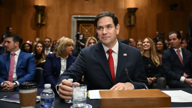 Rubio, in a suit and red tie, sitting at a table with a microphone and glass of water, with the committee room filled with seated people behind him