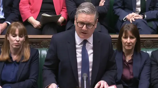 Keir Starmer stands at the despatch box in a black suit, white shirt and blue tie during PMQs. Sitting on the green leather benches behind him are Angela Rayner (L) in a black suit and blue shirt, her eyes partially closed, and Rachel Reeves (R) in a blue jacket and burgundy shirt