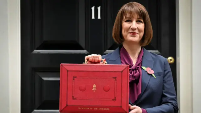 Rachel Reeves in dark suit, burgundy shirt with bow at the front and poppy pin on the left lapel holds up a ministerial red box ahead of the budget unveiling in front of 11 Downing Street