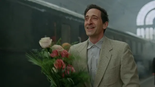 A still from The Brutalist shows Adrien Brody in a suit, holding a bunch of flowers and smiling