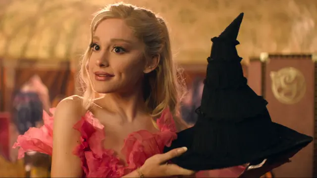 Ariana Grande, starring in Wicked, holding a witch's hat