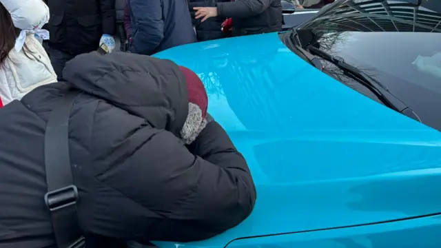 A woman covers her face and leans on a car crying