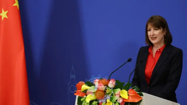 Chancellor Rachel Reeves in Beijing, China on Saturday. She is delivering a speech from a podium, with a mic and flowers in front of her. The backdrop is blue and a part of the flag of China (red with yellow stars) can be seen to the left of the photo.