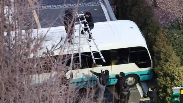 Two men standing on top of a bus, trying to hand over a ladder to people standing on the ground